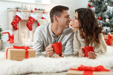 Happy couple with cups of hot drink on floor in room decorated for Christmas