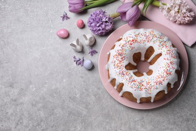 Glazed Easter cake with sprinkles, painted eggs and flowers on grey table, flat lay. Space for text