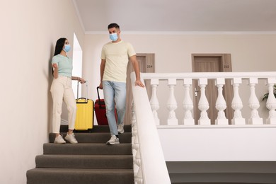 Couple wearing medical face masks with suitcases going down stairs in hotel