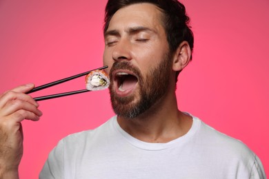 Handsome man eating tasty sushi roll with chopsticks on pink background