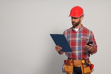 Professional builder in hard hat with clipboard, phone and tool belt on light background, space for text