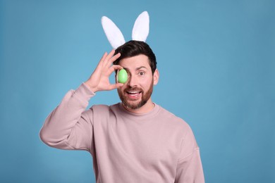 Photo of Happy man in cute bunny ears headband covering eye with Easter egg on light blue background