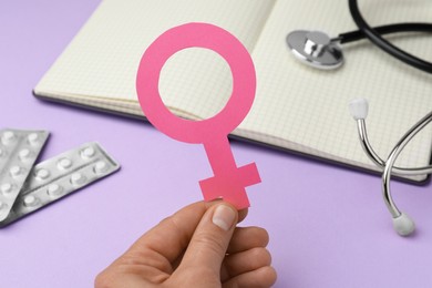 Photo of Girl holding female gender sign near open notebook, stethoscope and blisters of pills on violet background, closeup. Women's health concept