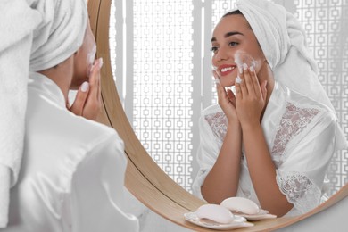 Beautiful young woman applying cleansing foam onto face near mirror in bathroom. Skin care cosmetic