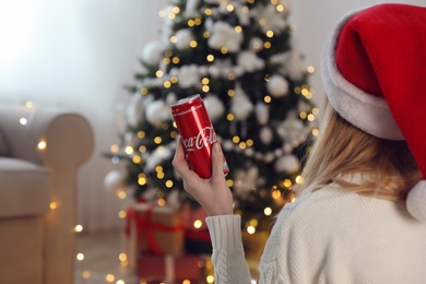 MYKOLAIV, UKRAINE - January 01, 2021: Woman in Santa hat with can of Coca-Cola against blurred Christmas tree at home, closeup
