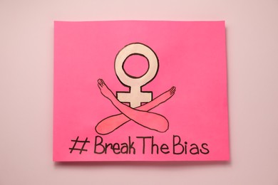 Card with hashtag BreakTheBias, gender female symbol and drawing of crossed arms on pink background, top view