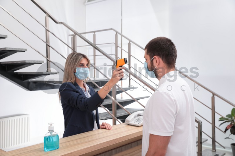Photo of Woman in mask measuring temperature of employee with noncontact thermometer at office reception