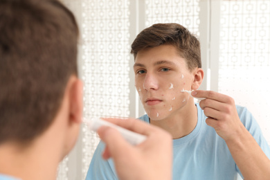 Teen guy with acne problem applying cream near mirror indoors