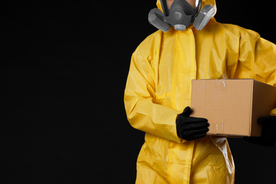 Man with cardboard box wearing chemical protective suit and space for text on black background, closeup. Prevention of virus spread
