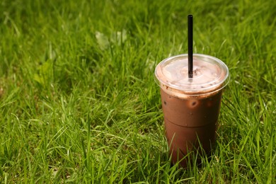 Takeaway plastic cup with cold coffee drink and straw on green grass outdoors, space for text