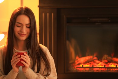 Young woman with cup of hot drink near fireplace indoors. Cozy atmosphere