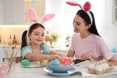 Happy mother with her cute daughter painting Easter eggs at table in kitchen