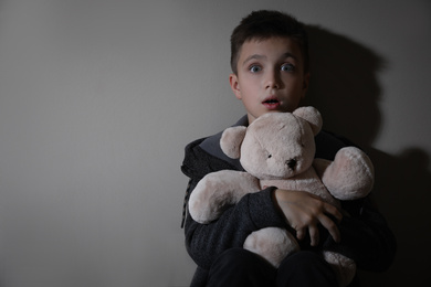 Photo of Scared little boy with teddy bear near beige wall, space for text. Domestic violence concept