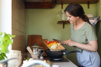 Young woman putting cut vegetables into frying pan in kitchen