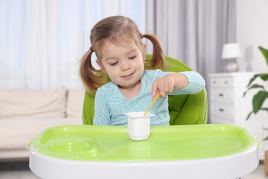 Photo of Cute little child eating tasty yogurt from plastic cup with spoon in high chair indoors