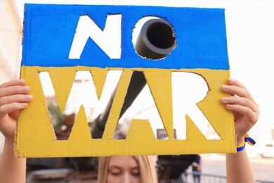 Woman holding poster in colors of Ukrainian flag with words No War near tank barrel on city street, closeup