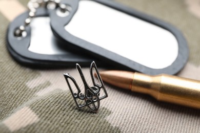 Ukrainian trident near military ID tags and bullet on camouflage background, closeup