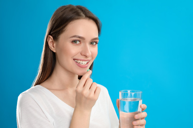 Young woman with glass of water taking vitamin pill on blue background
