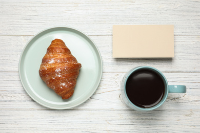 Croissant, coffee and blank card on white wooden table, flat lay. Tasty breakfast