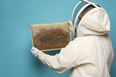 Beekeeper in uniform holding hive frame with honeycomb on light blue background, back view