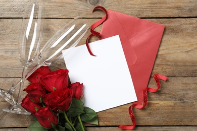 Flat lay composition with blank greeting card, glasses and roses on wooden table. Valentine's day celebration
