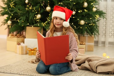 Photo of Cute little girl in Santa hat with book near tree and gifts at home. Christmas atmosphere
