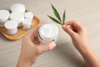 Woman holding jar of hemp cream and green leaf at wooden table, closeup. Natural cosmetics