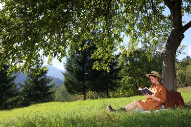 Young woman reading book under tree on meadow in mountains