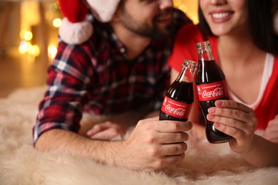 MYKOLAIV, UKRAINE - JANUARY 27, 2021: Young couple holding bottles of Coca-Cola in room with Christmas lights, closeup