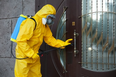 Person in hazmat suit disinfecting entrance door with sprayer. Surface treatment during coronavirus pandemic