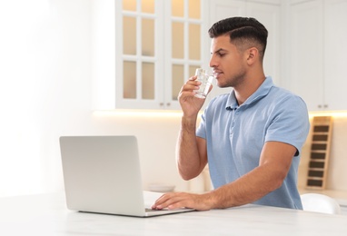 Man drinking pure water from glass while working with laptop in kitchen