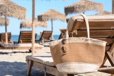 Photo of Straw bag with sunglasses on wooden sunbed near sea. Beach accessories