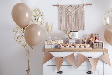 Baby shower party. Different delicious treats on white wooden chest of drawers and decor near light wall