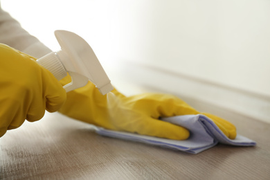 Person in gloves cleaning wooden table with spray detergent and rag indoors, closeup