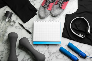 Flat lay composition with sportswear, notebook and dumbbells on white marble background. Gym workout plan