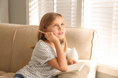 Thoughtful little girl with pencil and notebook on couch at home