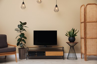 Modern TV on cabinet, armchair and green plants near beige wall in room. Interior design