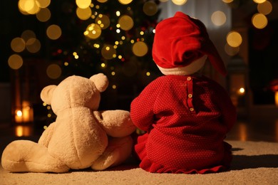 Baby wearing Santa hat with teddy bear in room decorated for Christmas, back view