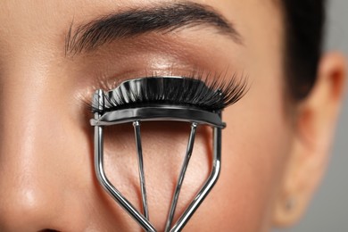 Young woman using eyelash curler on grey background, closeup