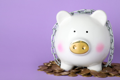 Piggy bank  with steel chain and coins and lilac background, space for text. Money safety concept