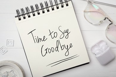 Notebook with phrase Time to say Goodbye, eyeglasses and earphones on white wooden table, flat lay