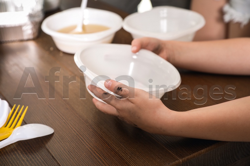 Poor little child holding empty plate over wooden table in charity centre, closeup