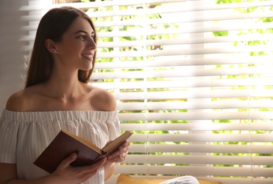 Photo of Beautiful young woman reading book near window at home