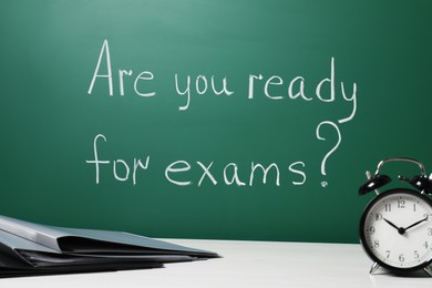 Office folders and alarm clock on white table near chalkboard with phrase Are You Ready For Exams