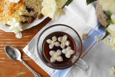 Photo of Glass cup of aromatic tea with beautiful jasmine flowers and dessert on wooden table, flat lay