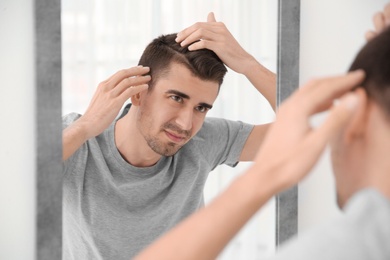 Young man with hair loss problem looking in mirror indoors