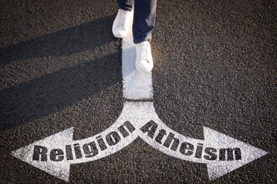 Image of Choice between atheism and religion. Man walking towards drawn marks on road, closeup. Arrows with words pointing in opposite directions