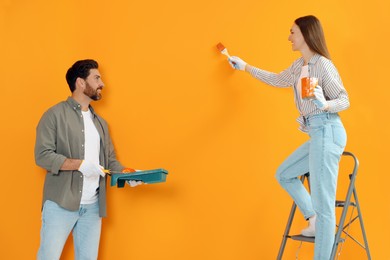 Photo of Woman painting orange wall and man holding container with roller. Interior design