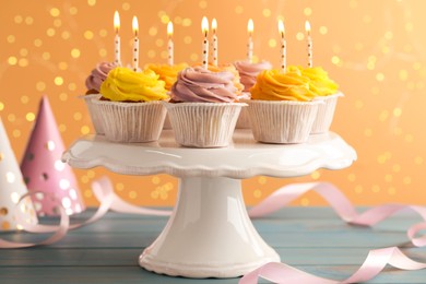 White stand with tasty birthday cupcakes on light blue wooden table against blurred lights