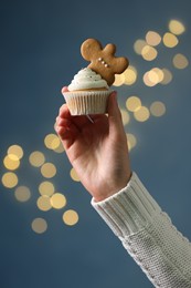 Photo of Woman holding tasty Christmas cupcake with gingerbread man cookie against blurred festive lights, closeup